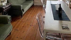 Une pièce avec un canapé vert, un plancher en bois, une table à manger avec des chaises, une bouteille de vin et une vue dégagée sur l’extérieur grâce à une grande fenêtre.