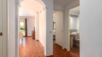 Um corredor com piso ladrilhado, uma porta que dá para um banheiro e vista para um jardim por uma porta aberta.