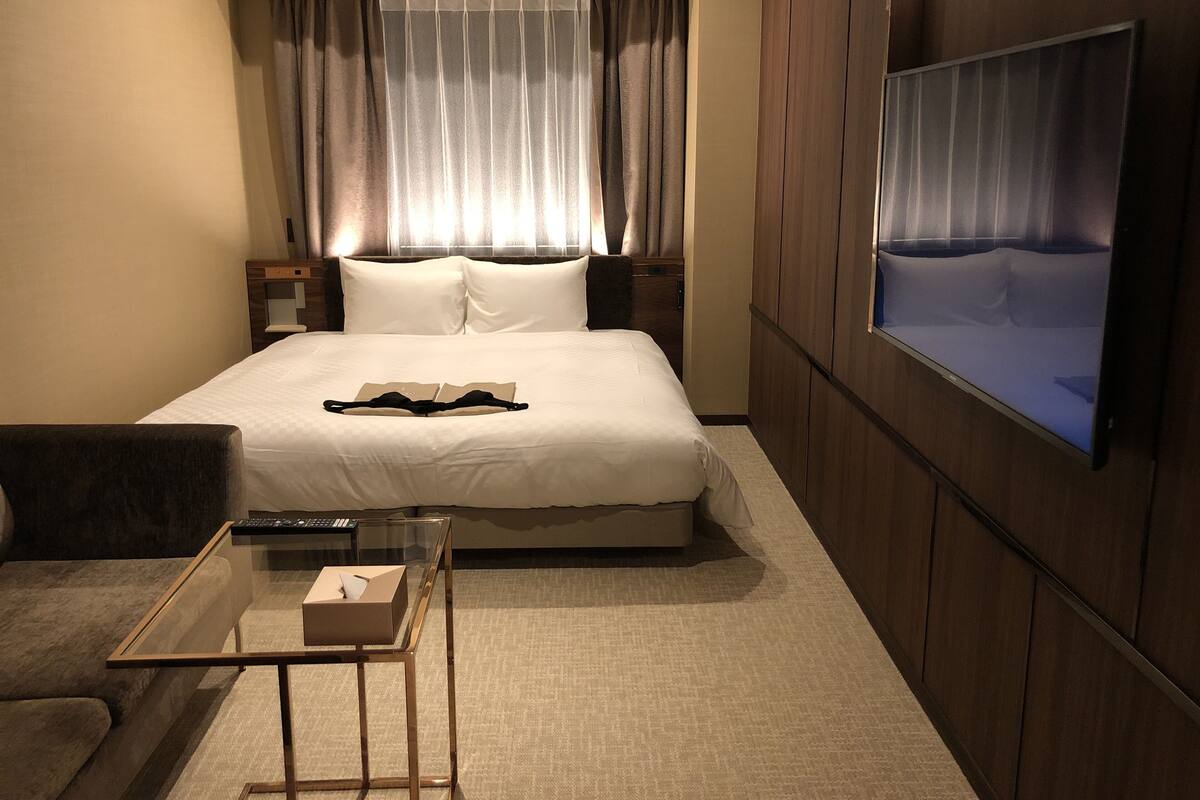 Una habitación de hotel con una cama grande, un televisor en la pared, una mesita de noche con un control remoto y una pequeña mesa con una caja.