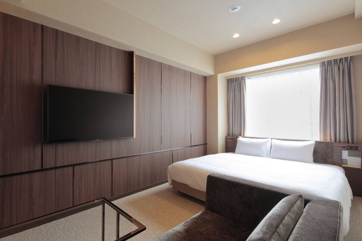 Habitación de hotel con una cama grande, un televisor de pantalla plana montado en la pared, una ventana con cortinas y un pequeño rincón de descanso con un sofá.