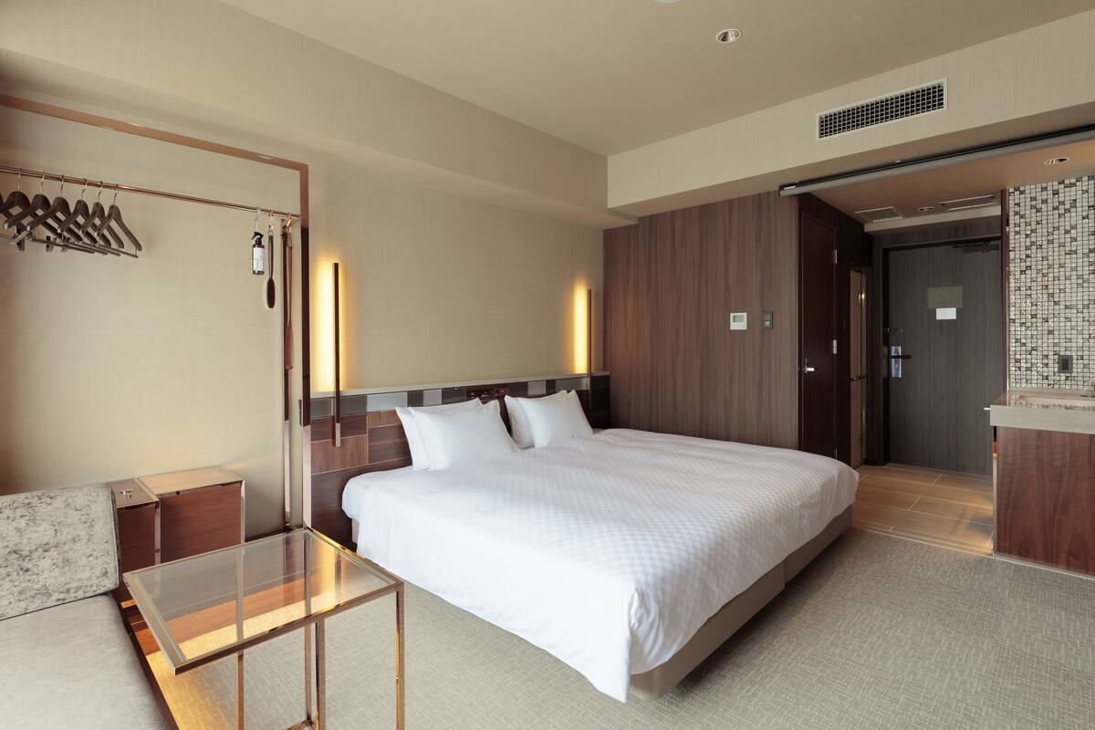 Habitación de hotel moderna con una cama grande, una mesa de vidrio, un armario y un baño visible a través de una puerta abierta.