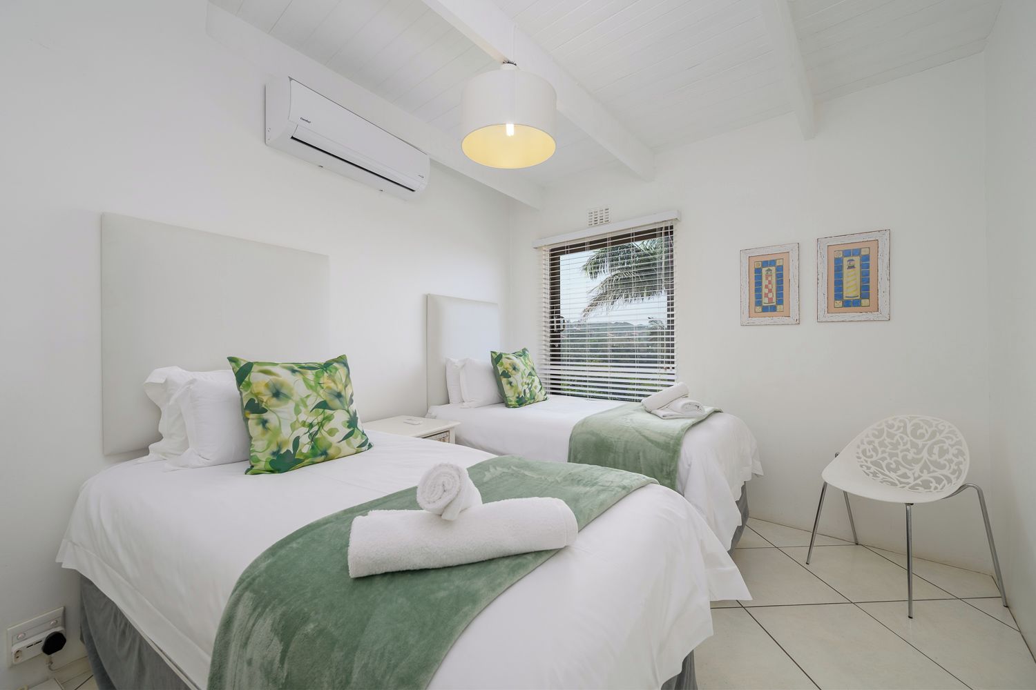 A hotel room with two beds, a chair, a window with blinds, and a wall-mounted air conditioner.