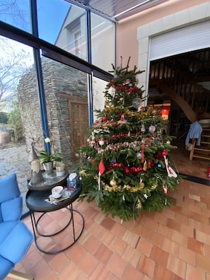 A decorated Christmas tree in a room with a stone wall, a blue chair, and a table with a cup and saucer.