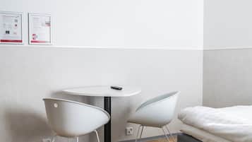A small, modern room with a round table and two white chairs, a bed with a white duvet, and two informational posters on the wall.