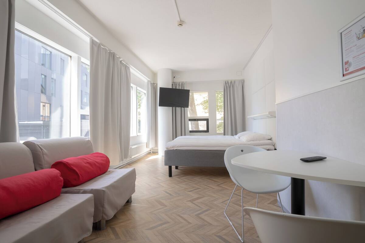A modern hotel room with a bed, a sofa with red cushions, a small table, and a TV mounted on the wall.