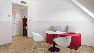 Une petite pièce moderne aux couleurs blanc et rouge, comprenant une table ronde, deux chaises blanches, un canapé rouge avec un coussin gris et un parquet en bois à motifs chevrons.