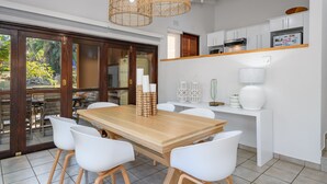Une cuisine moderne avec des meubles blancs, une table à manger en bois et des chaises blanches. Des suspensions en osier sont accrochées au-dessus de la table.