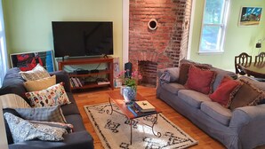 Un salon avec un canapé, une table basse sur laquelle sont posés des livres et un vase, un téléviseur sur un meuble, un mur en briques et une fenêtre.