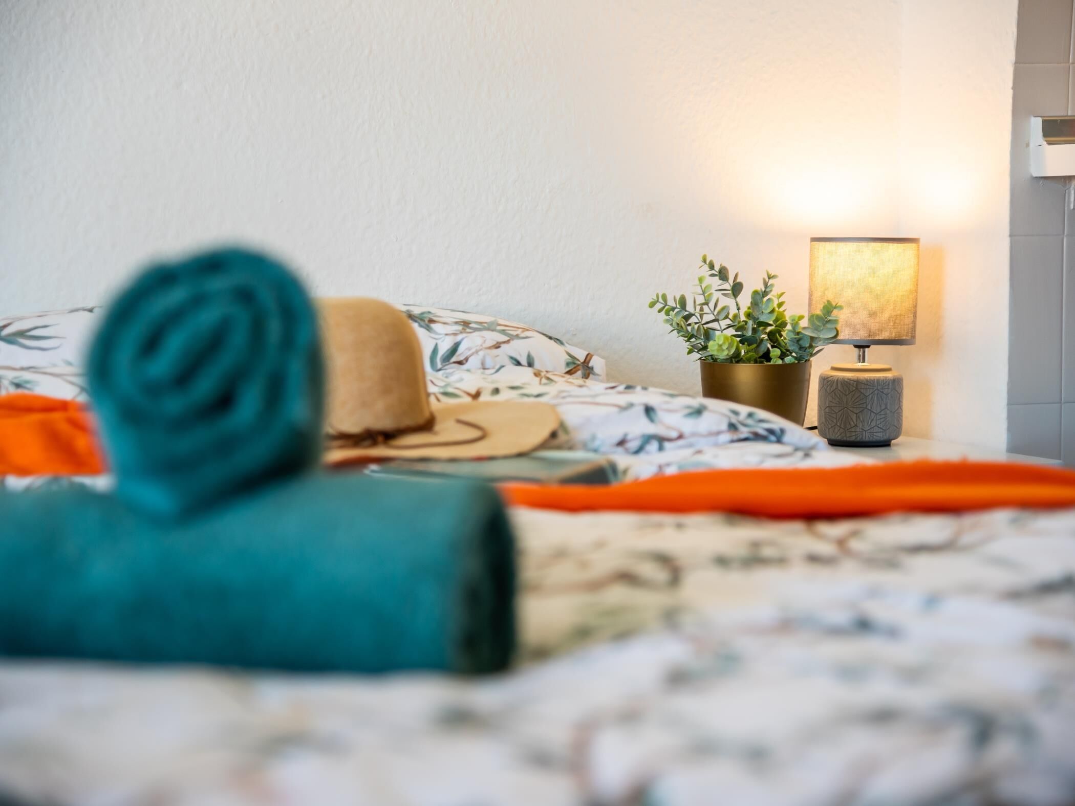Una cama bien tendida con una colcha de estampado floral, una toalla verde azulada enrollada y un sombrero color beige sobre las almohadas. Una pequeña planta en maceta y una lámpara de noche están sobre la mesita junto a la cama.