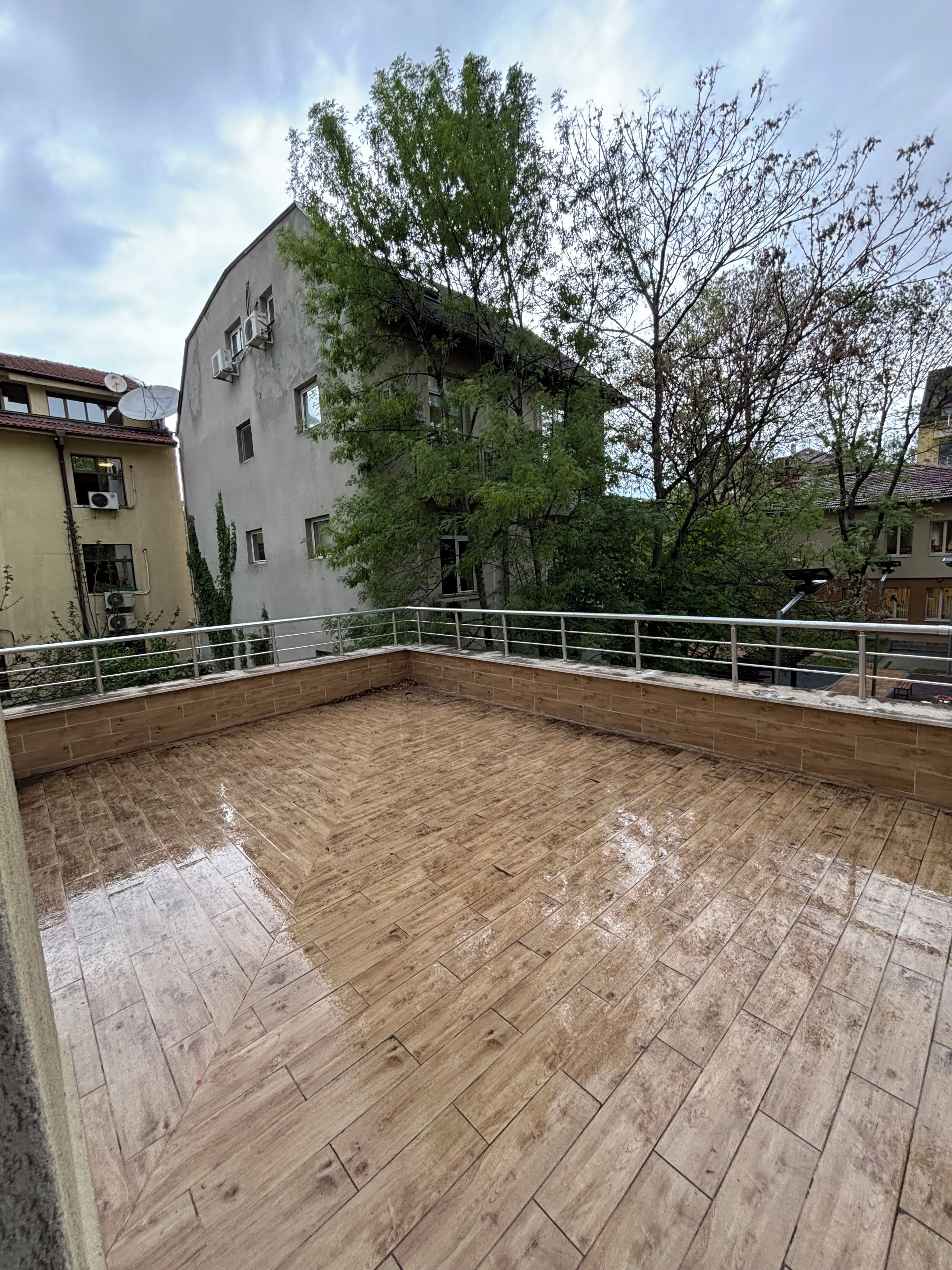 A rooftop terrace with a wooden floor, surrounded by a metal railing, and buildings in the background.