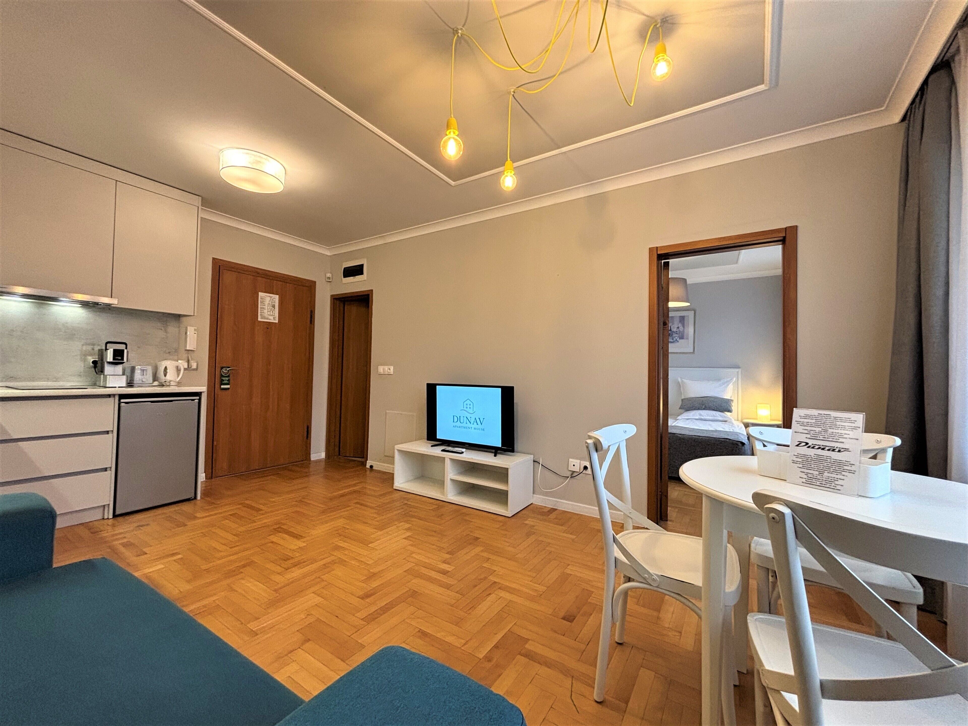A compact hotel room with a kitchenette, a flat-screen TV, a dining area with a table and chairs, and a bed with a headboard.