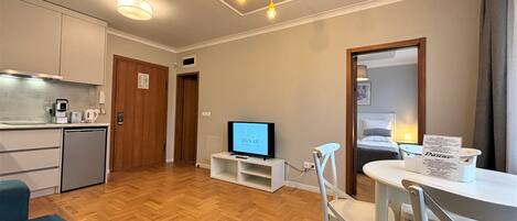 A compact hotel room with a kitchenette, a flat-screen TV, a dining area with a table and chairs, and a bed with a headboard.