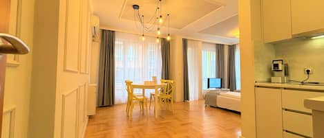A modern hotel room with a wooden herringbone floor, a dining area with a table and chairs, a bed, a TV, and a kitchenette.