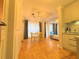 A modern hotel room with a wooden herringbone floor, a dining area with a table and chairs, a bed, a TV, and a kitchenette.