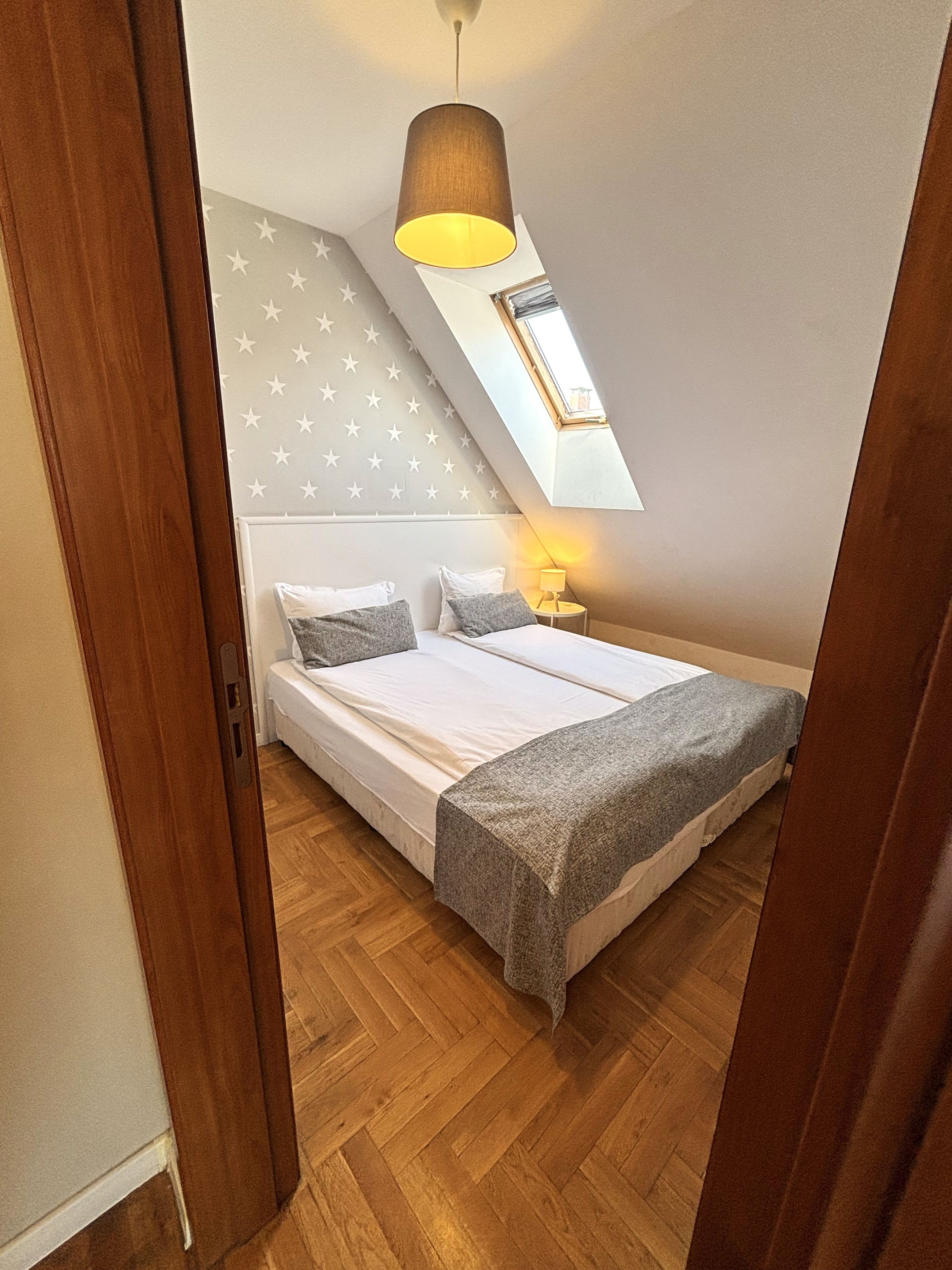 A bedroom with a bed, a nightstand, a lamp, a star-patterned wall, and a sloped ceiling with a skylight.