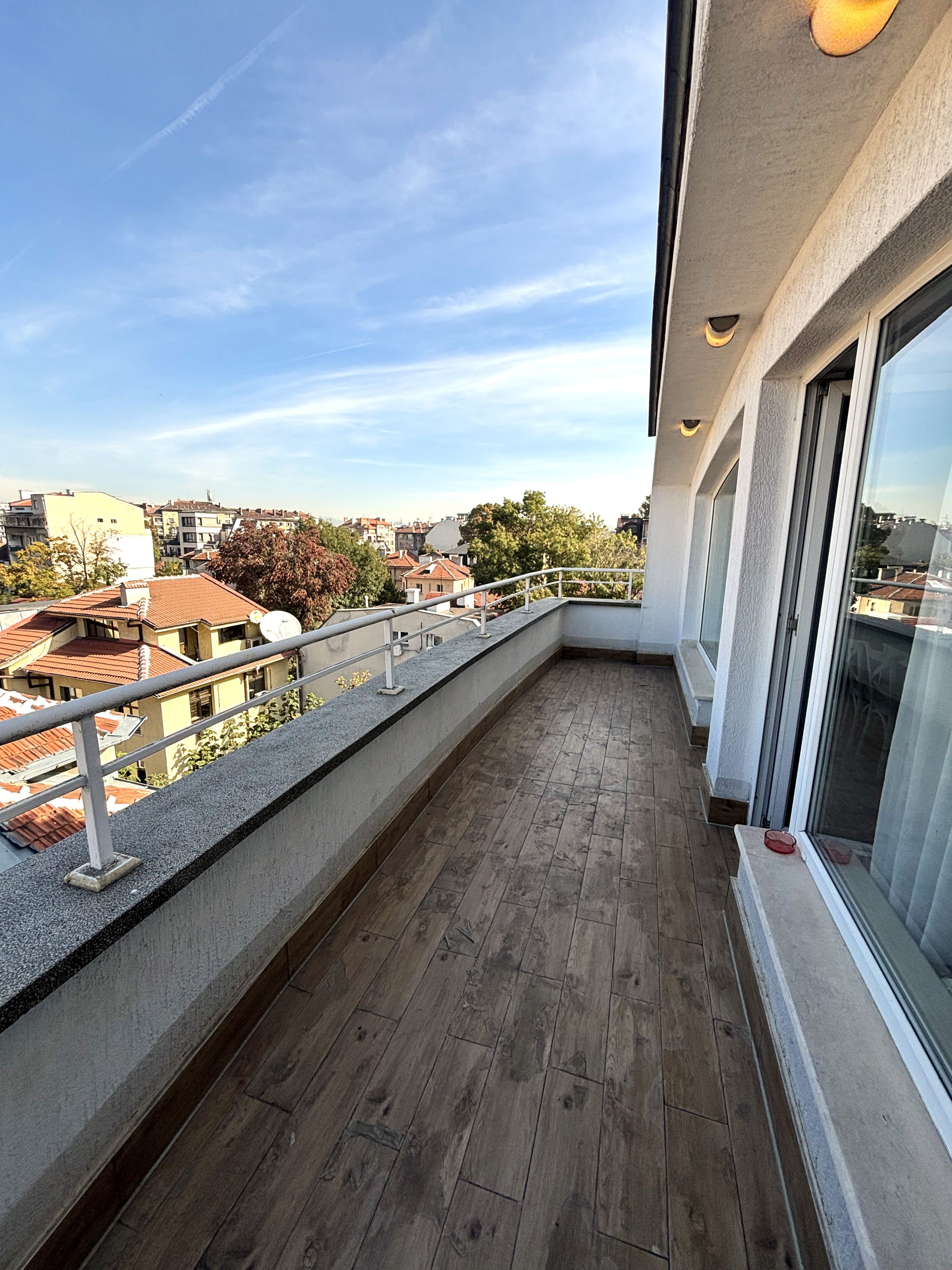 Un balcón con piso de madera, barandilla de metal y amplios ventanales que ofrecen vistas de la ciudad.