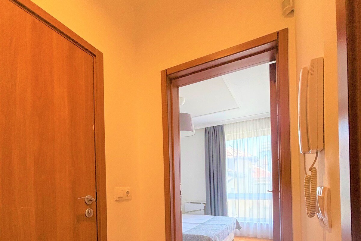 A hallway leading to a bedroom with a bed, a telephone, and a window with curtains.