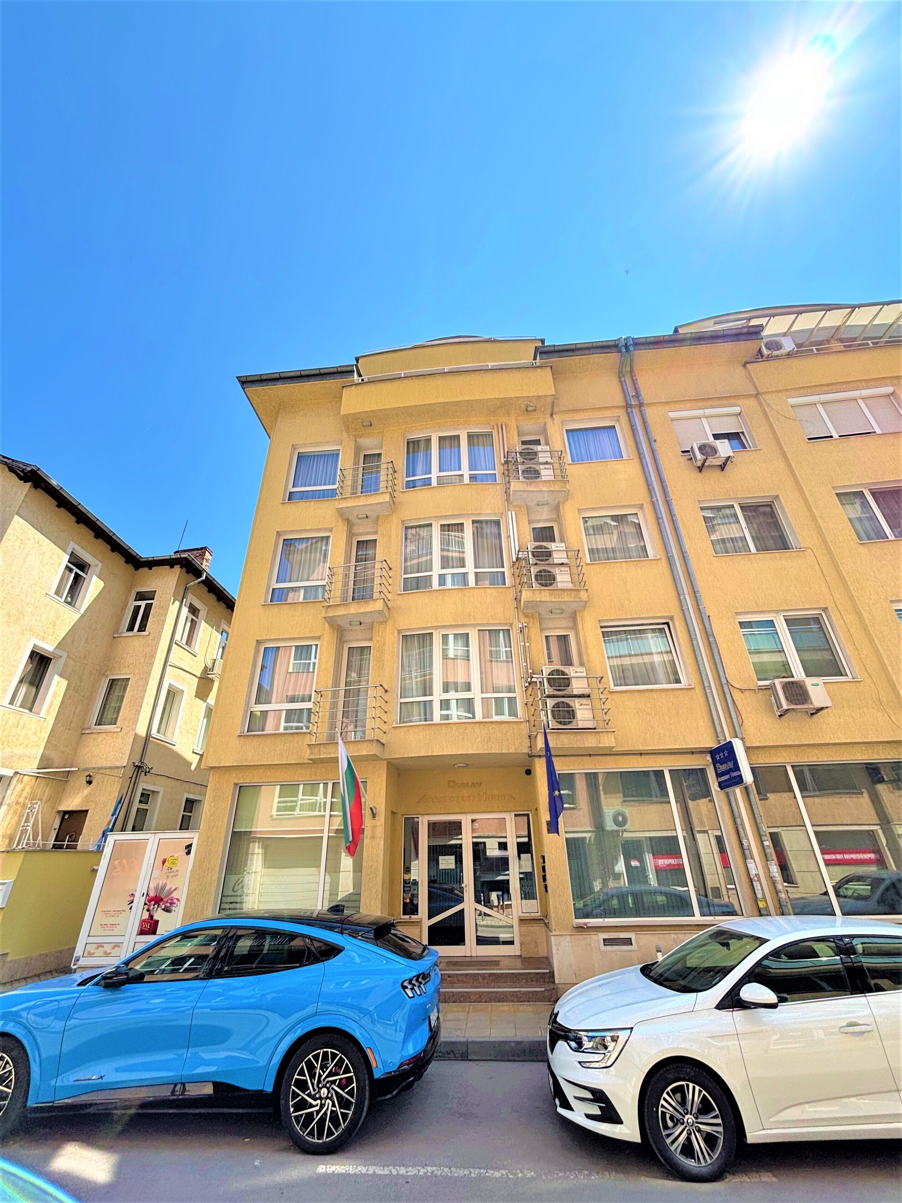 A multi-story building with a yellow facade, multiple windows, and balconies. There are two cars parked in front, and a flag is visible on the left side.