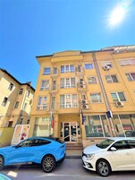 A multi-story building with a yellow facade, multiple windows, and balconies. There are two cars parked in front, and a flag is visible on the left side.