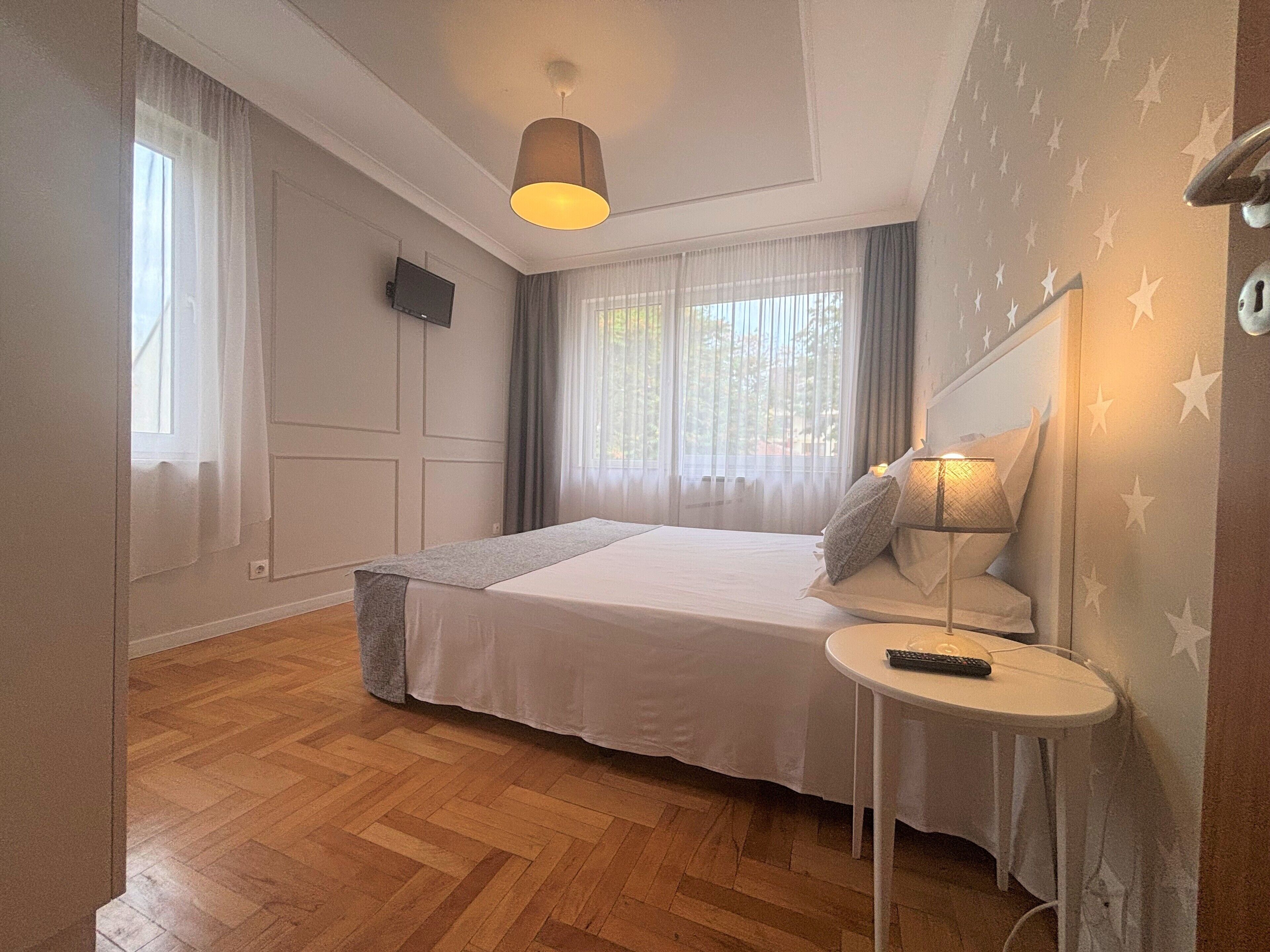 Un dormitorio con cama, mesita de noche, lámpara, televisor y ventana con cortinas.