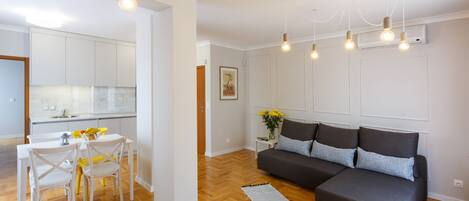 A modern living room with a grey sectional sofa, a dining table with chairs, and a kitchen area with white cabinets.