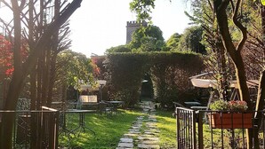 Ein Gartenweg führt zu einer Sitzgelegenheit mit Tischen und Stühlen, umgeben von Grünpflanzen und einer dahinterliegenden Turmstruktur.