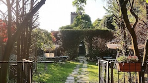 A garden pathway leading to a seating area with tables and chairs, surrounded by greenery and a tower structure in the background.