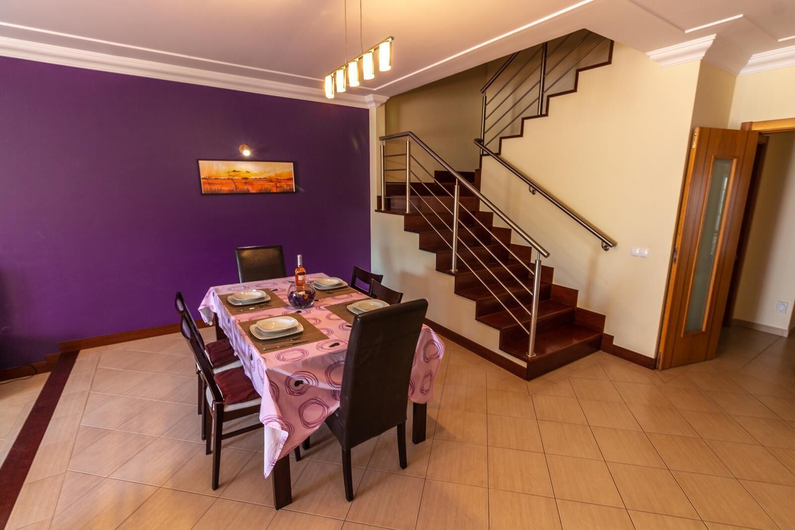 A dining area with a table set for four, a staircase, and a purple wall with a painting.