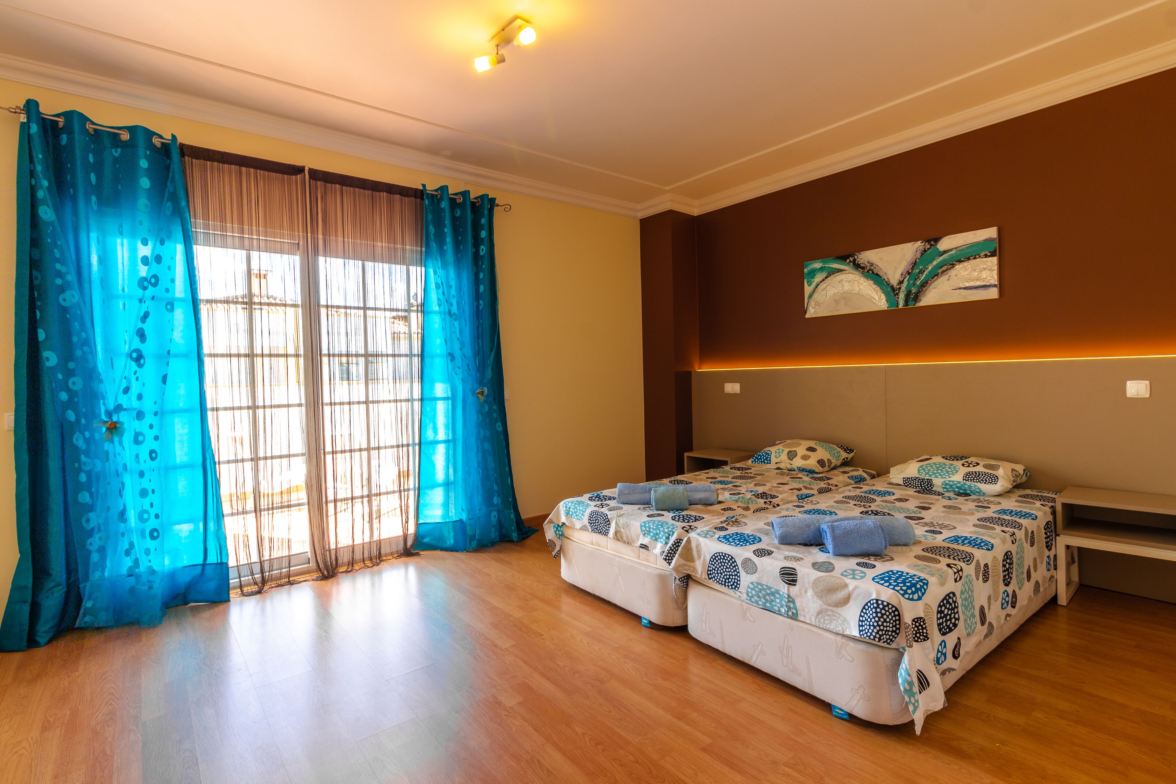 Un dormitorio con dos camas, un ventanal con cortinas, un cuadro en la pared y un banco.