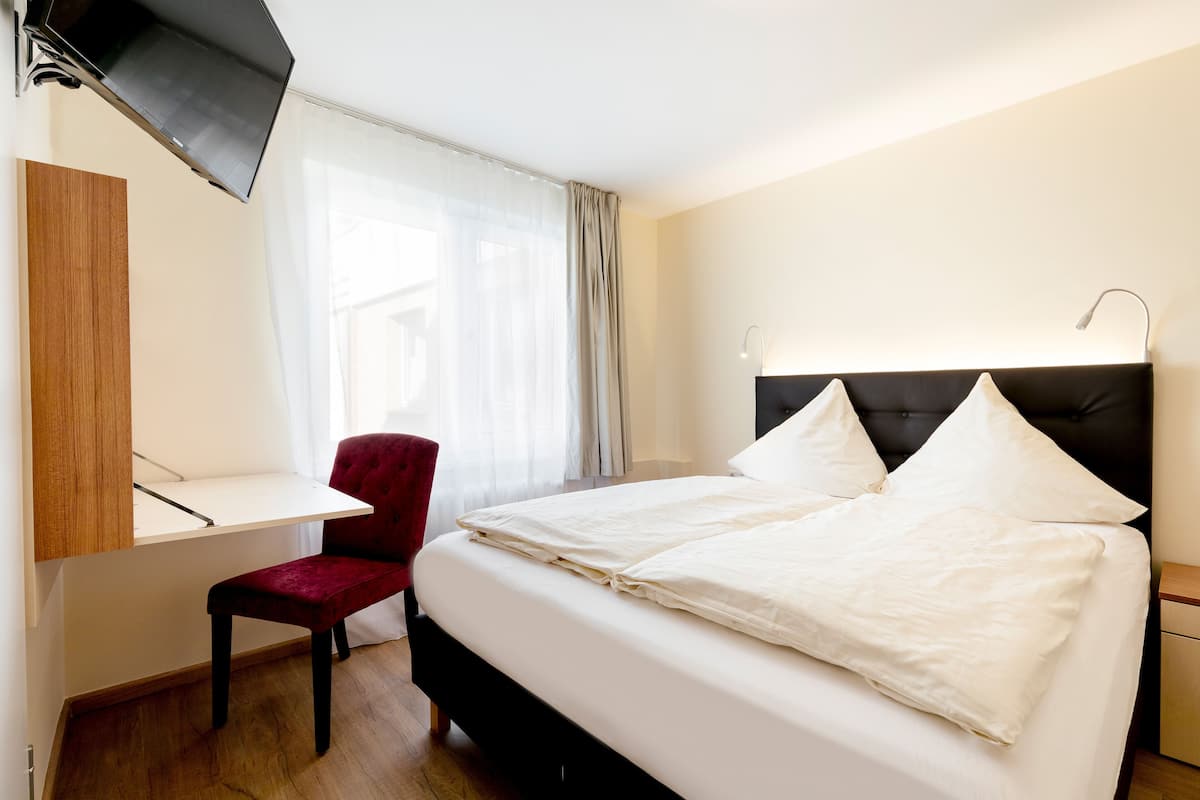 Quarto de hotel com cama, escrivaninha, cadeira, televisão e janela com cortinas.
