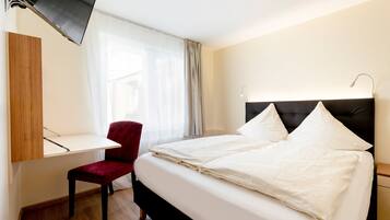 Quarto de hotel com cama, escrivaninha, cadeira, televisão e janela com cortinas.