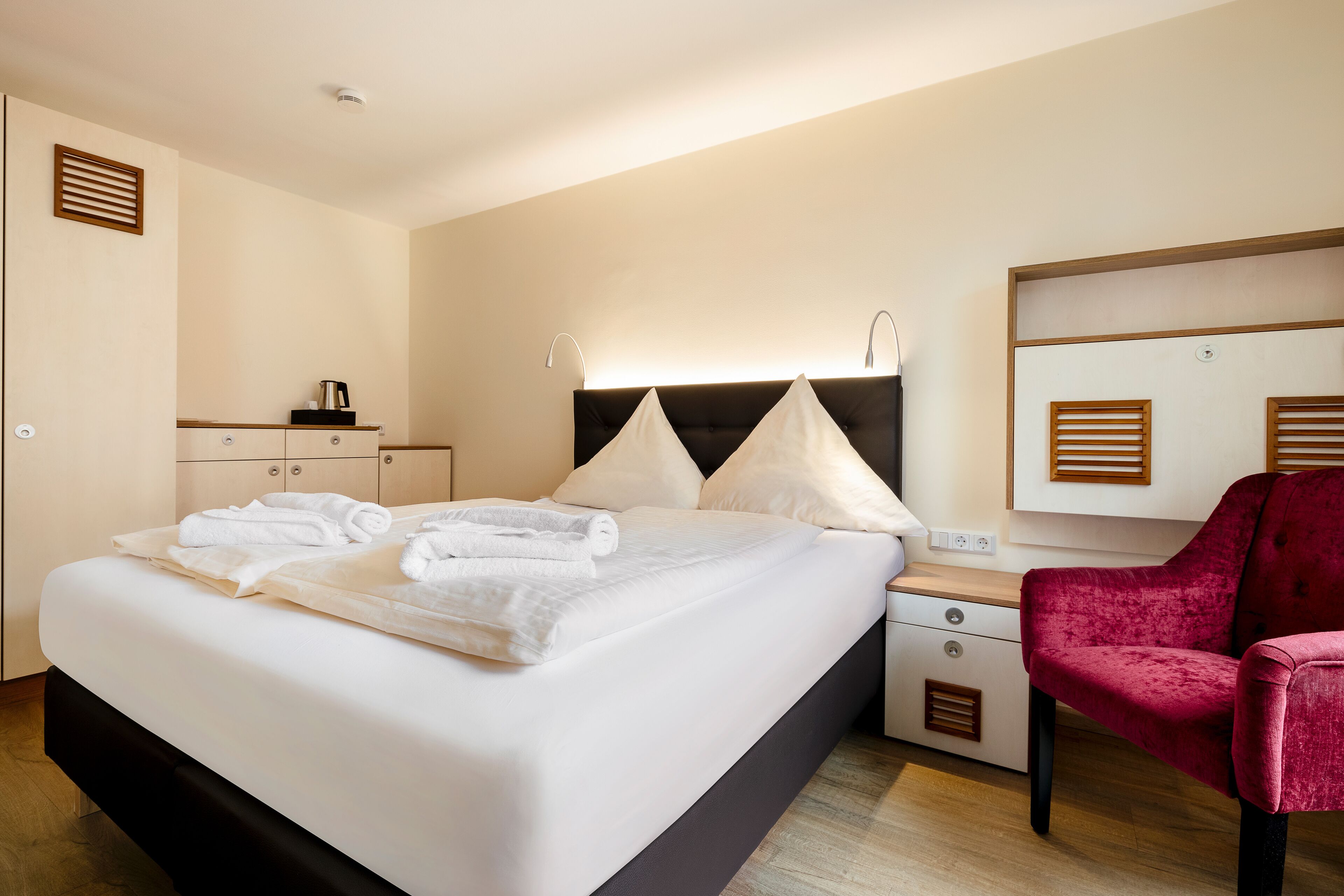 Habitación de hotel con una cama bien hecha, una silla roja, una mesita de noche con una lámpara y un espejo sobre la cama.
