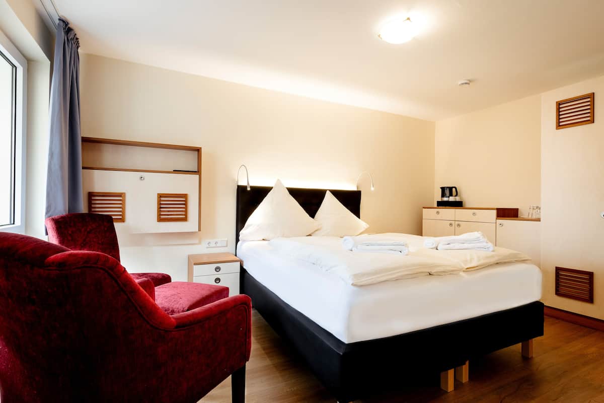 Quarto de hotel com cama, uma cadeira vermelha, mesa de cabeceira, escrivaninha e guarda-roupa.