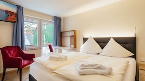 A hotel room with a large bed, a red chair, a desk, and a window with curtains. - Hotel Helgolandia (Helgoland)