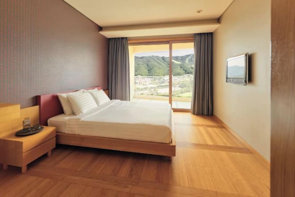 A modern hotel room with a large bed, wooden flooring, a nightstand, a TV, and a view of a mountainous landscape through the window.