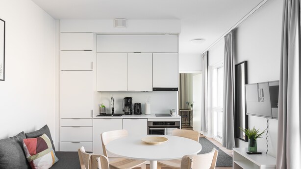 Een moderne, goed verlichte woonruimte met een ronde eettafel, lichte stoelen, een grijze bank met kleurrijke kussens, een keuken op de achtergrond en een groot raam met gordijnen.