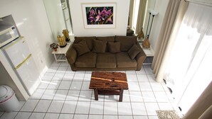 Ein Wohnzimmer mit einem braunen Sofa, einem Holztisch, einem gefliesten Boden in Weiß, einem gerahmten Blumenbild an der Wand und einem weißen Kühlschrank.