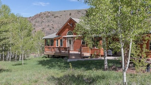 A log cabin with a porch and a deck, surrounded by trees and a hill in the background. - Taylormade Getaway - Almont (Almont)