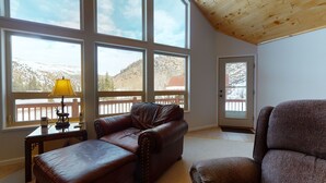 A spacious living room with large windows, a leather sofa, a patterned armchair, and a wooden ceiling. - Taylormade Getaway - Almont (Almont)