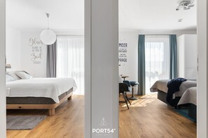 A modern hotel room with a wooden floor, a bed with a white duvet, a desk with a chair, and a window with curtains. - Onkel Olli - Infrared sauna, fireplace, balcony, beach chair, water view, (Olpenitz)