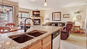 A kitchen with granite countertops, a double sink, and a dishwasher. There is a dining area with a table and chairs, and a living room with a sofa and a fireplace. - 1 Bedroom Ski in Condo Only One Block From Historic Downtown, Shopping, Restaurants and Entertainment (Breckenridge)