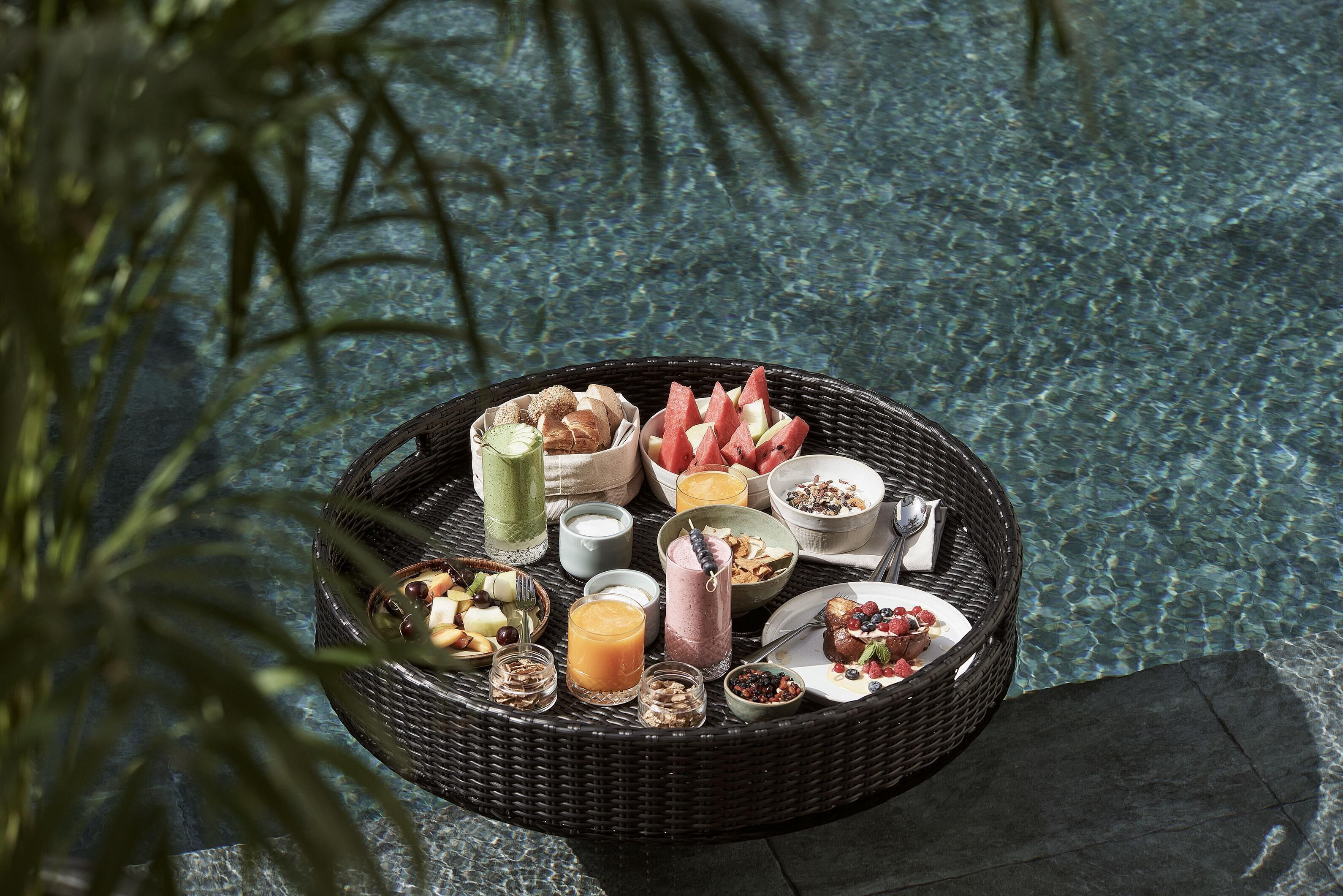 A breakfast buffet with various food items and beverages on a wicker table by a pool.