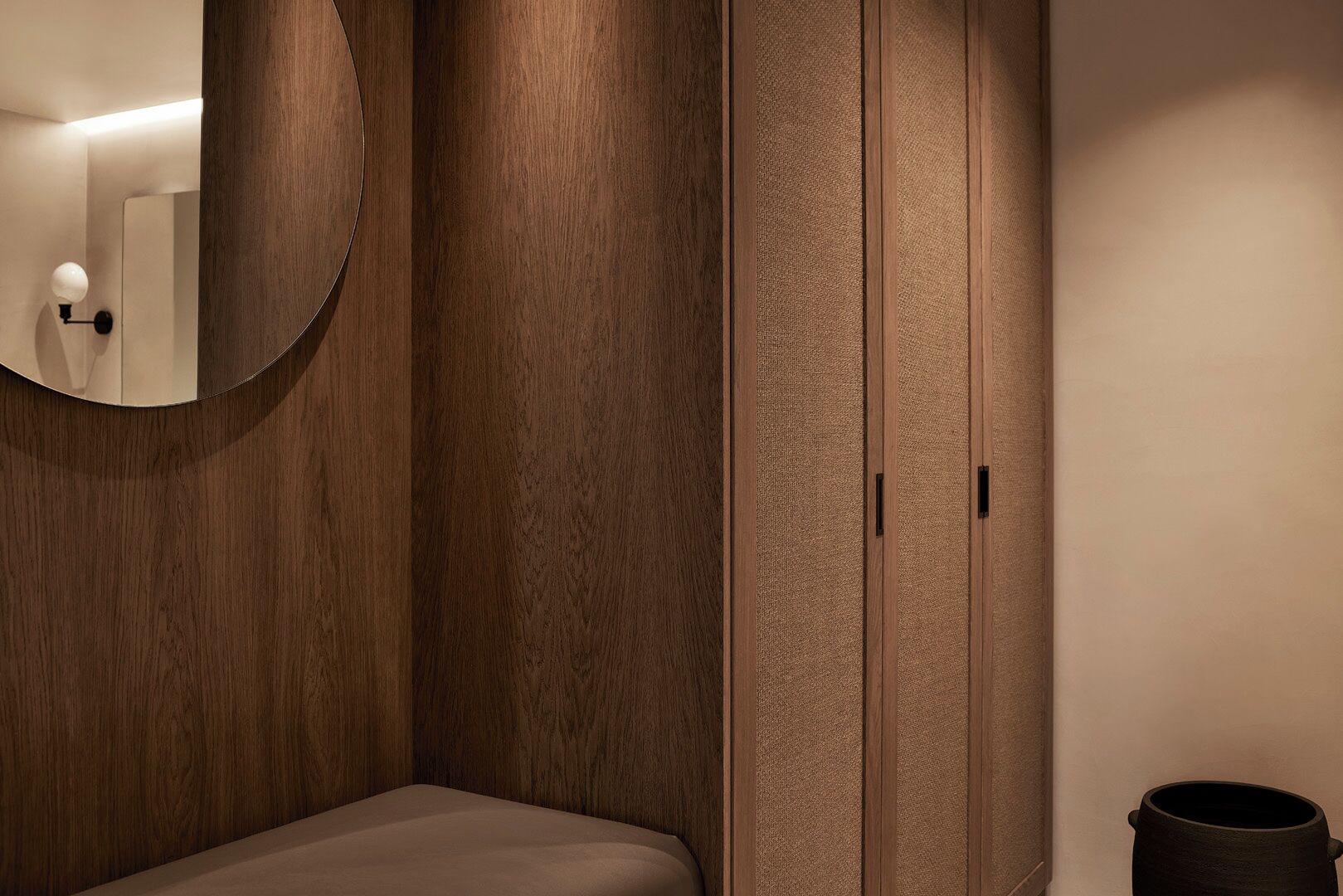 A bedroom with a wooden wardrobe, a round mirror, and a small trash bin.
