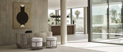 A modern lobby with a reception desk, two white cushioned stools, a large abstract artwork, and a view of the outdoors through large windows.