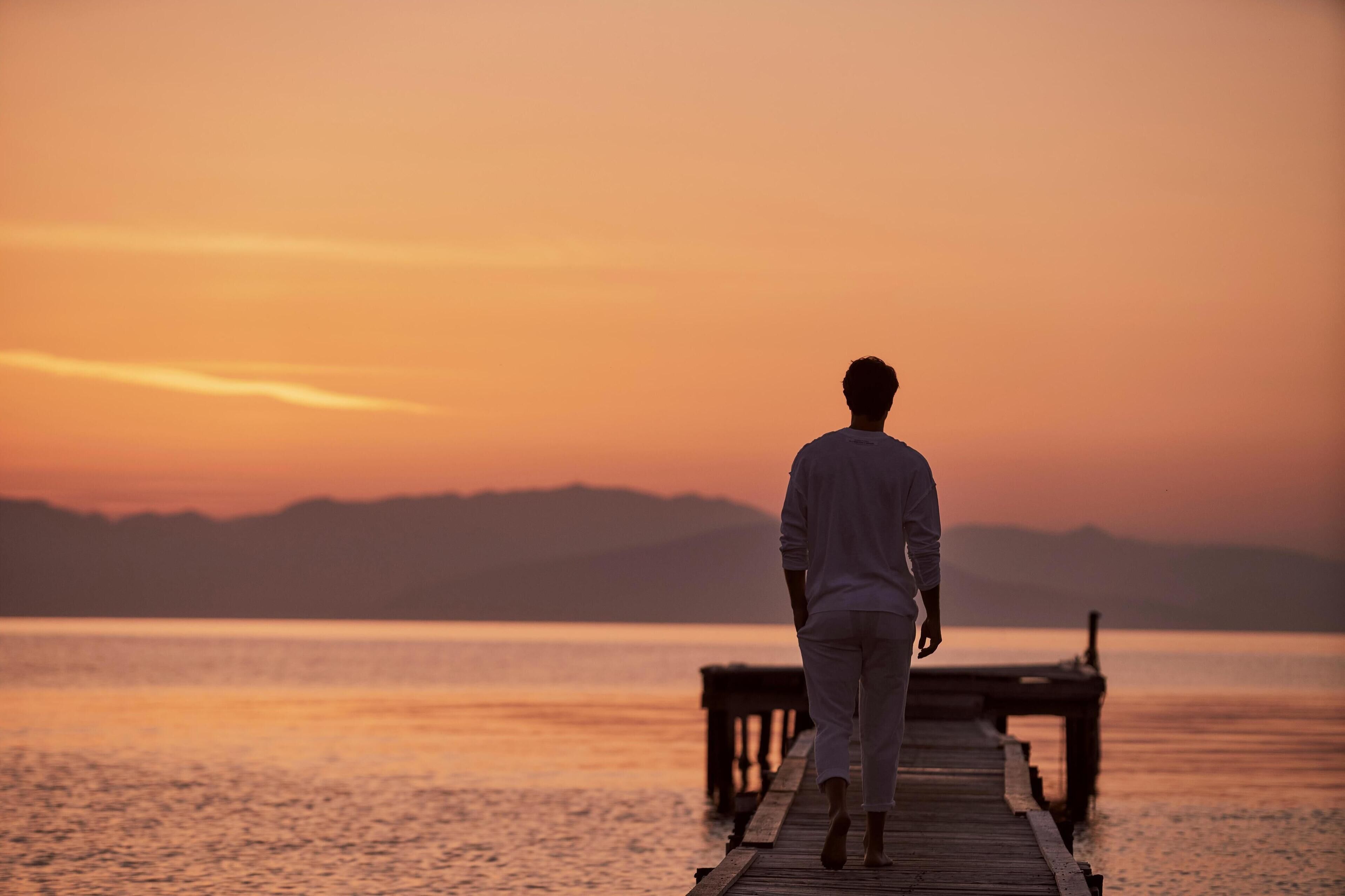 A person walking on a wooden pier extending into a calm body of water during sunset.