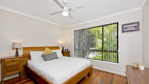 Ein Schlafzimmer mit einem hölzernen Bett, einem Nachttisch, einer Lampe, einem Deckenventilator, einem Fenster mit Jalousien und einem gerahmten Bild an der Wand.