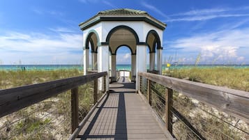 A wooden boardwalk leading to a white gazebo with green accents and arches, overlooking a beach with clear turquoise water.