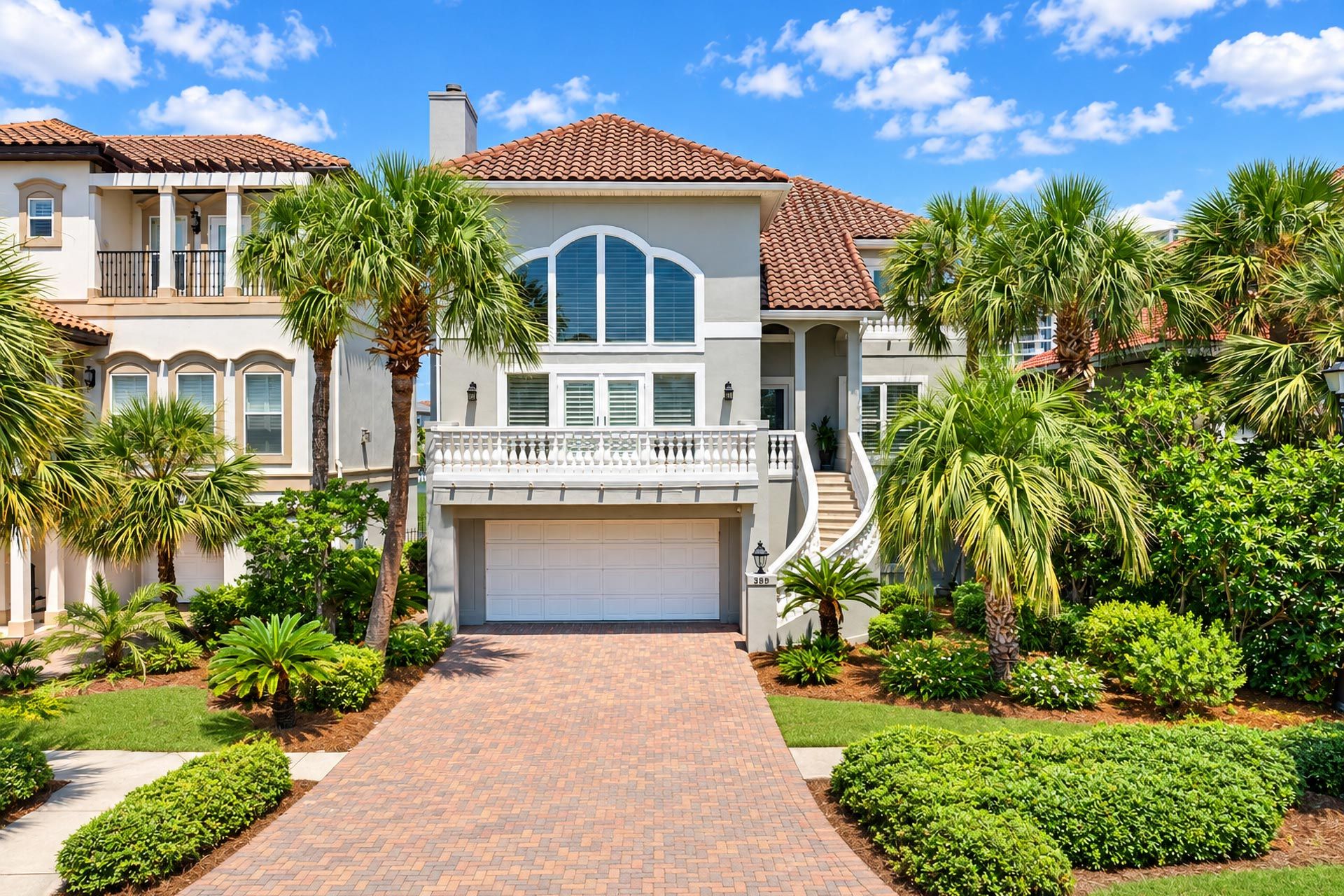 A two-story house with a brick driveway, a garage, and a well-maintained garden with palm trees and shrubs.