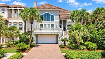 A two-story house with a brick driveway, a garage, and a well-maintained garden with palm trees and shrubs.