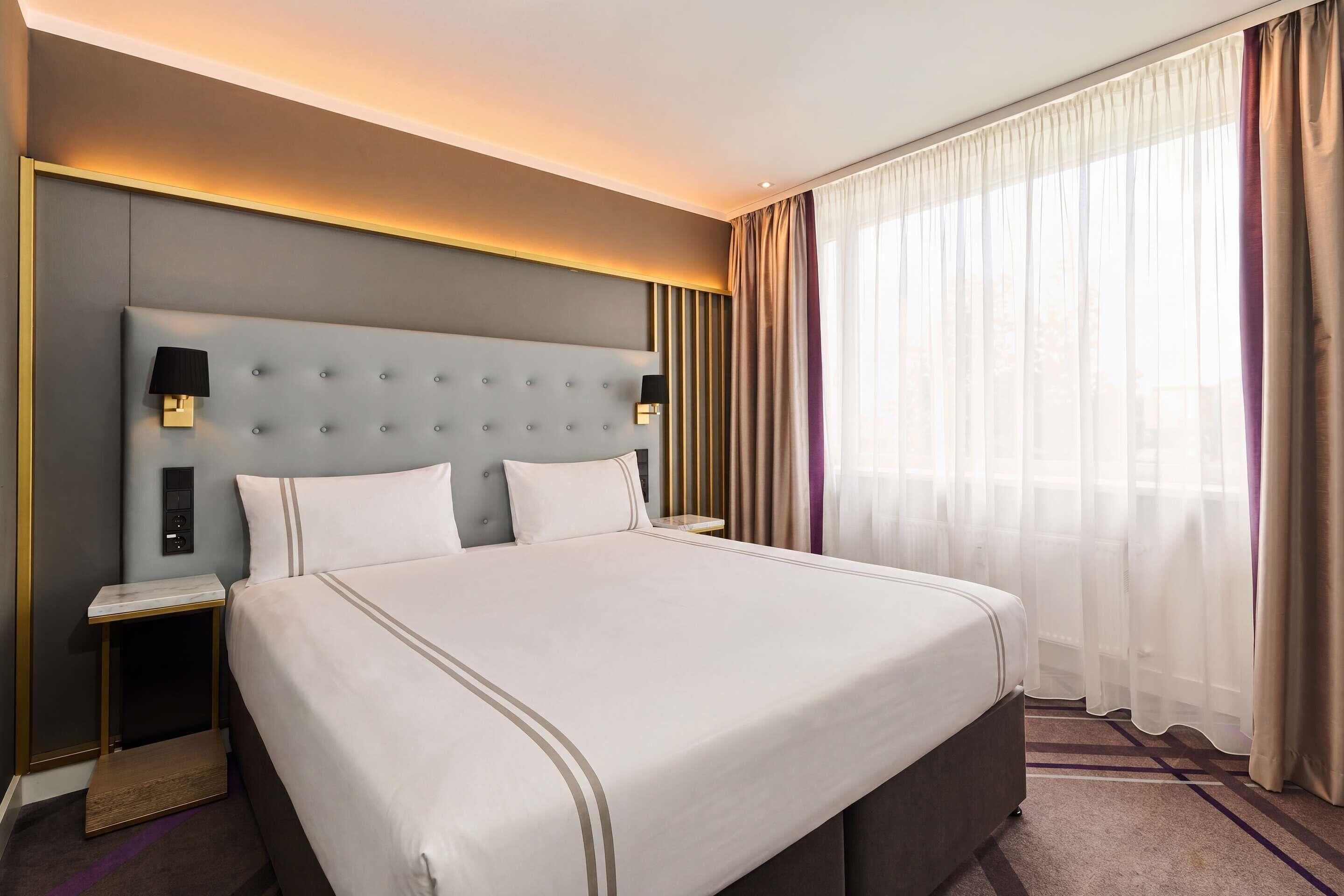Ein Hotelzimmer mit einem großen Bett, einem Nachttisch, einer Wandleuchte und einem Fenster mit transparenten Vorhängen.
